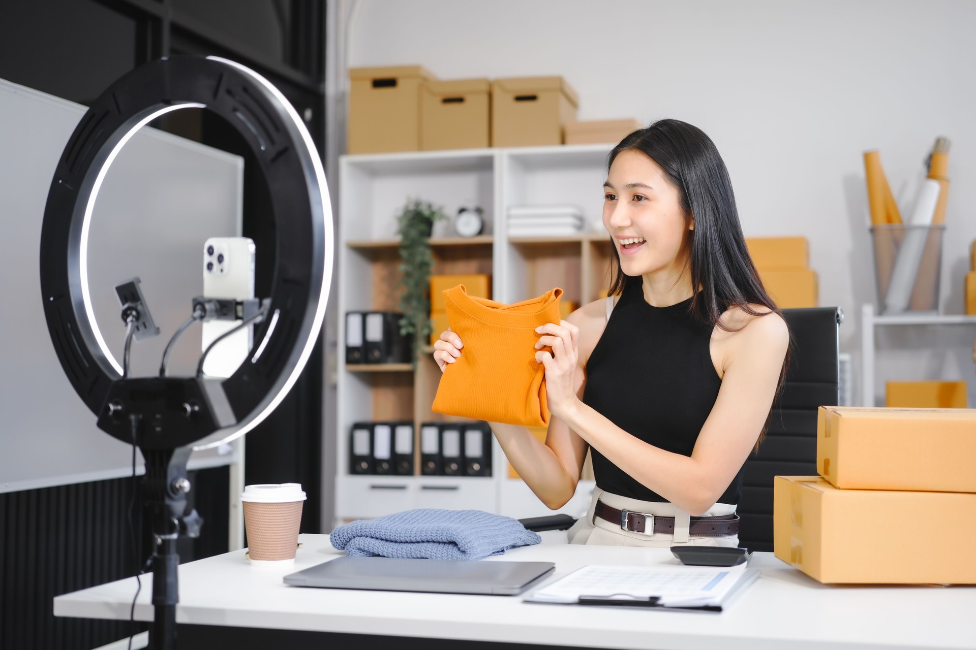 An Asian female business owner records a live vlog, showing a parcel box in front of a smartphone. She sells clothes online, engaging customers and promoting products for successful e-commerce sales