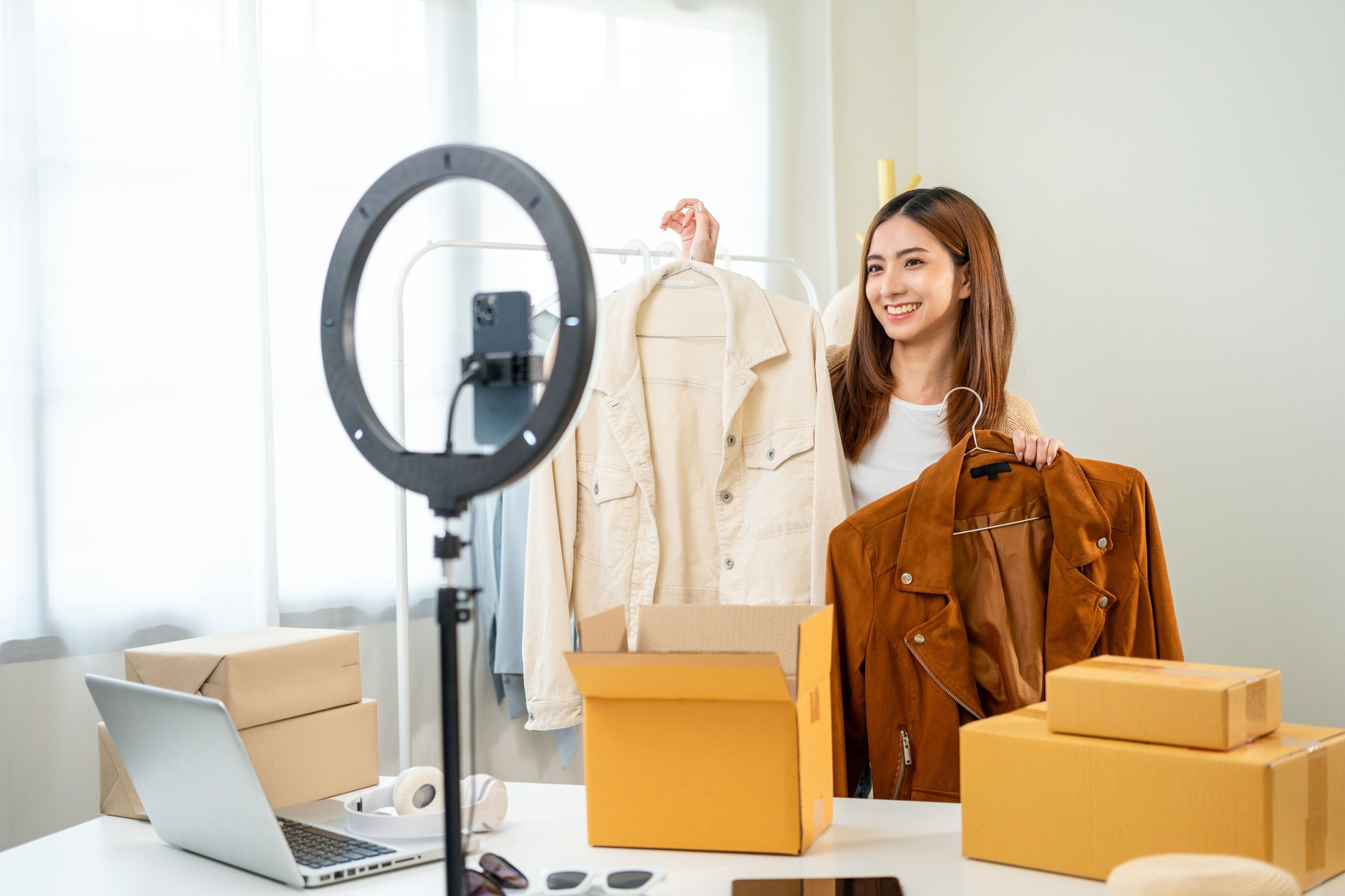 Young asian woman business owner at fashion store using smartphone live streaming for sale fashion clothing. Influencers record video review clothes for sell on social media. Online shopping concept.